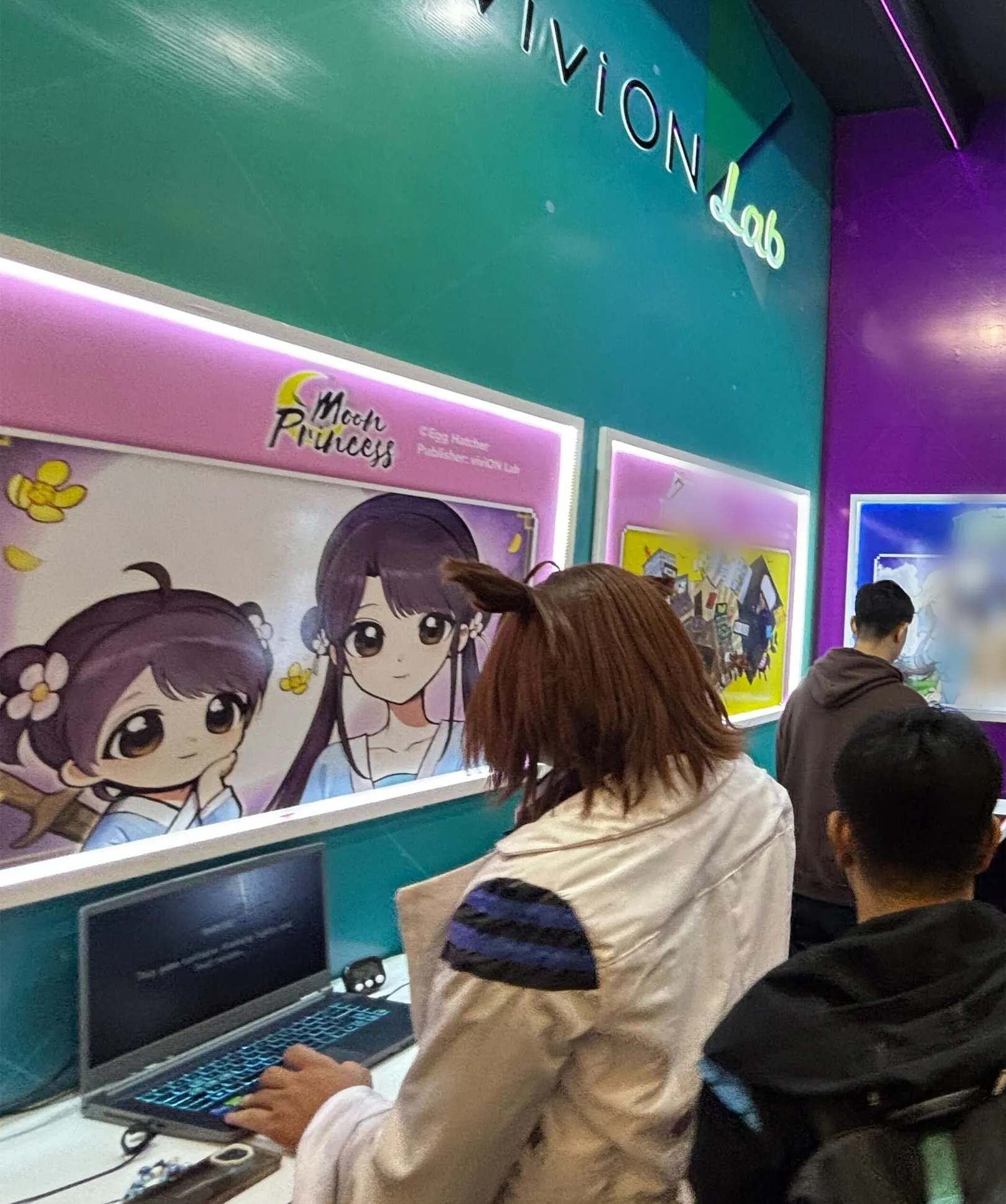 A visitor playing Moon Princess at a laptop, below an illuminated panel of the game's key art.